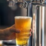 A pint of beer held in a hand inside a pub, showing golden ale and a frothy head.